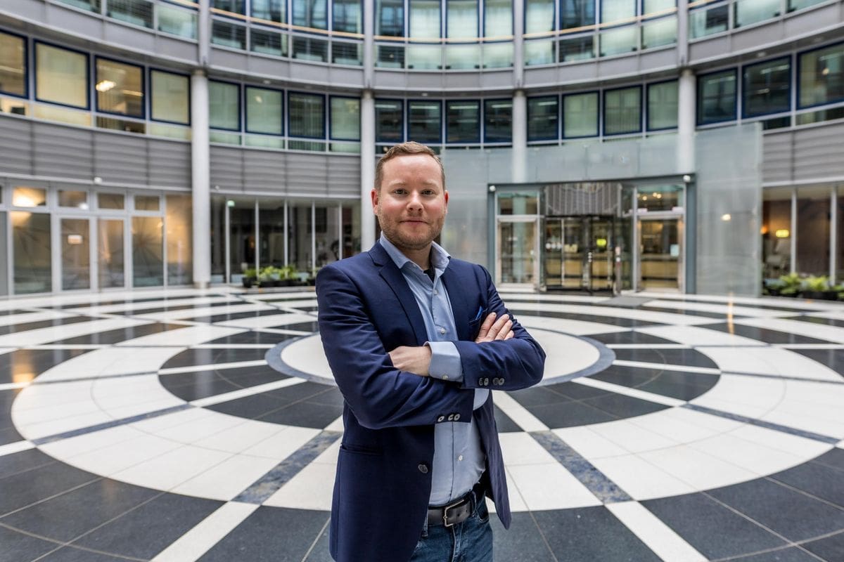 Ein Mann mit kurzem Haar und blauem Sakko steht mit verschränkten Armen in der Mitte eines runden, modern gestalteten Atriums mit schwarz-weißem Boden.
