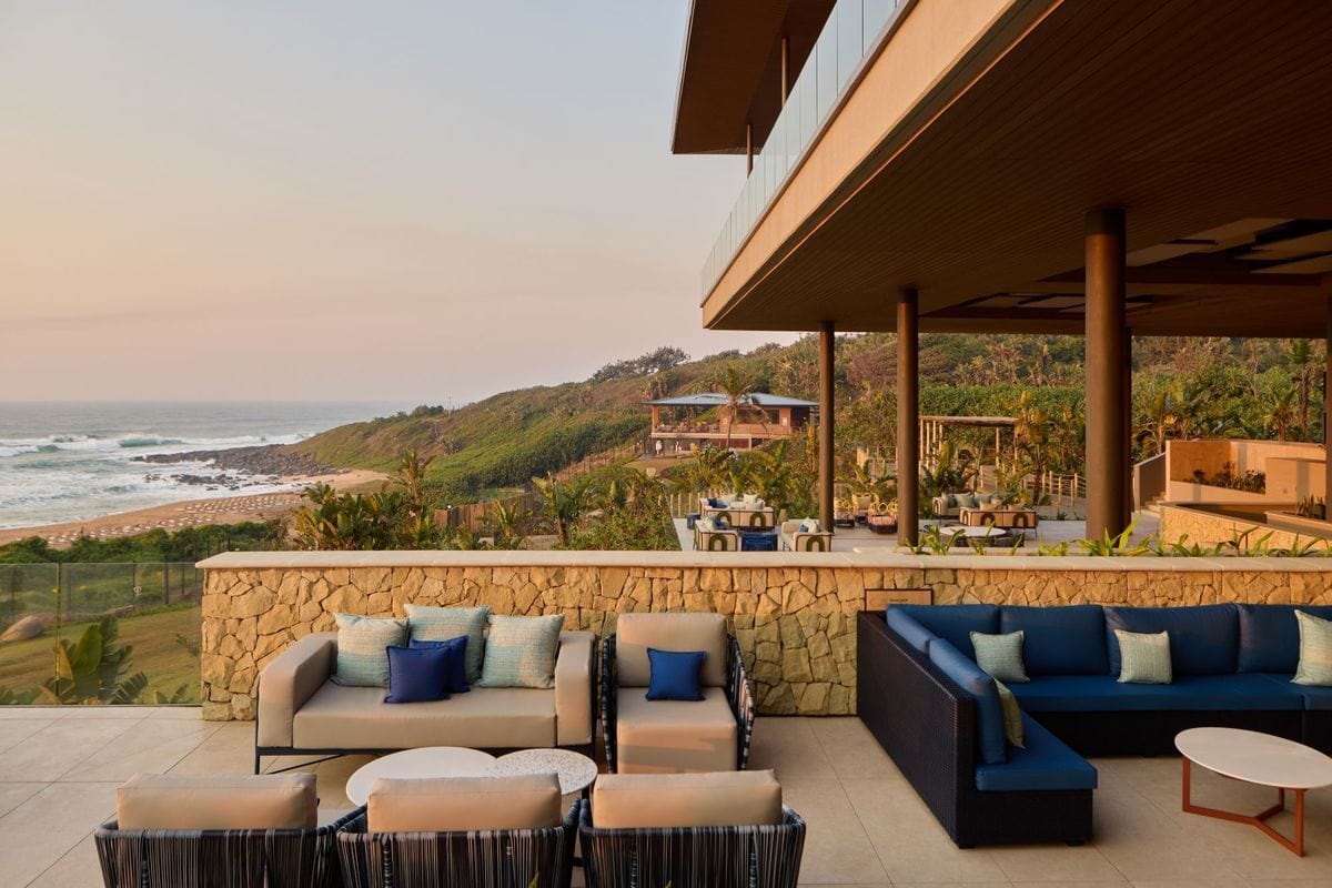 Eine großzügige Hotelterrasse mit bequemen Loungemöbeln und Blick auf eine hügelige Küstenlandschaft mit Sandstrand und dem Ozean bei sanftem Abendlicht.