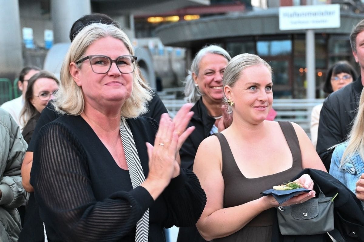 Eine blonde Frau mit Brille klatscht in die Hände, daneben hält eine junge Frau mit braunem Top einen kleinen Teller, im Hintergrund weitere Personen.