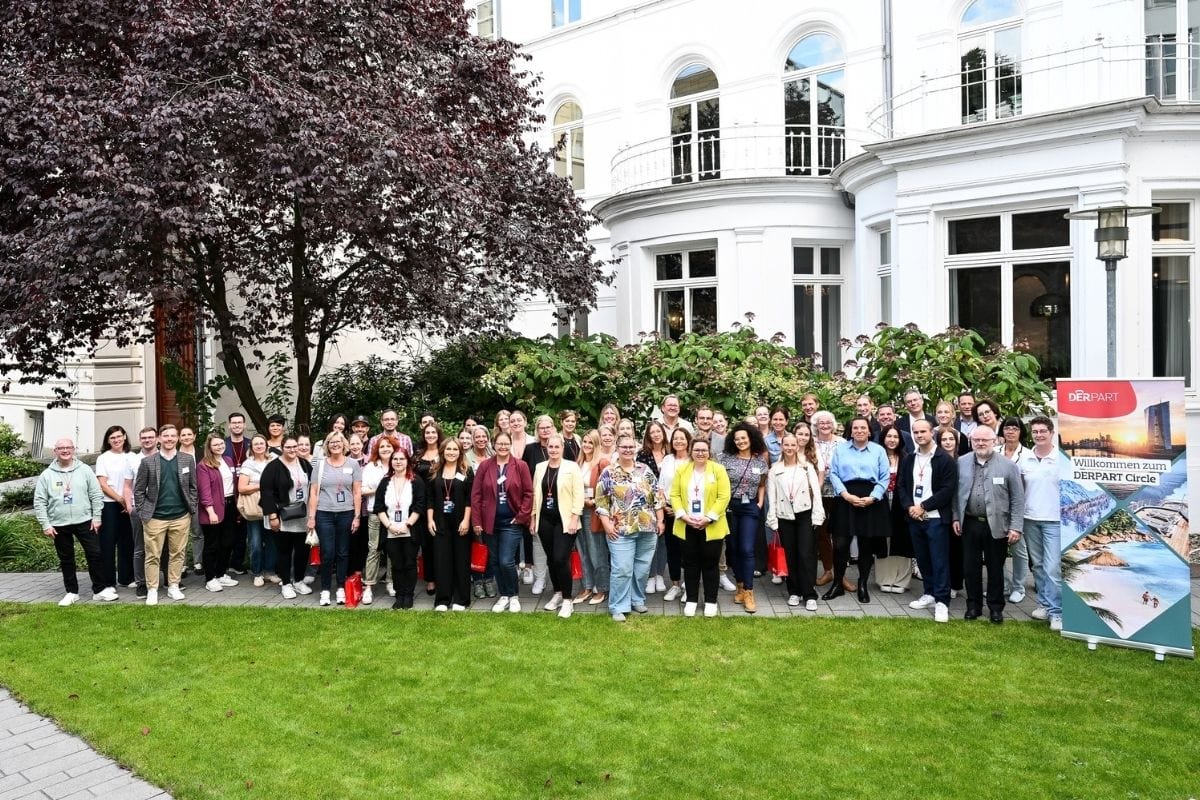 Eine große Gruppe von Menschen steht in einer Reihe vor einem weißen Gebäude und einem Baum auf einer Rasenfläche, rechts steht ein DERPART-Aufsteller.