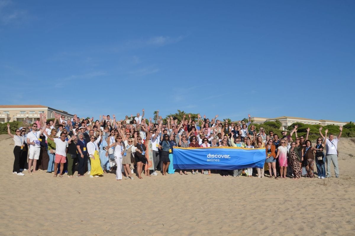 Eine große Gruppe von Menschen steht an einem sonnigen Sandstrand und hält gemeinsam ein blaues Banner mit der Aufschrift discover. airlines in die Höhe.