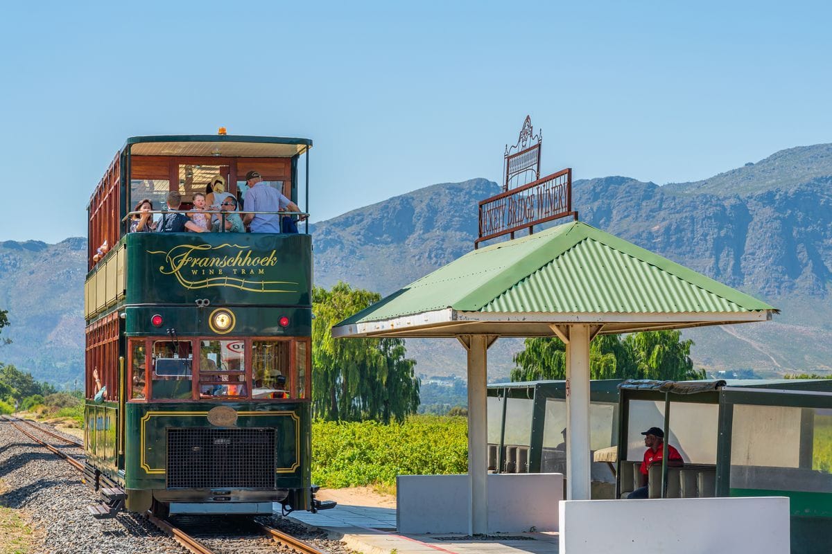 Eine grüne, doppelstöckige Franschhoek Wine Tram hält an einer kleinen Haltestelle vor einer Bergkulisse, während Fahrgäste auf dem Oberdeck sitzen.