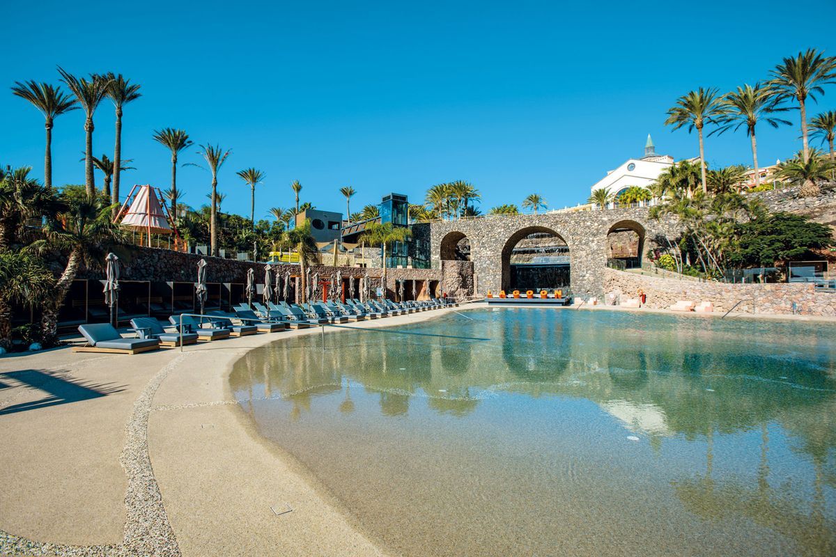 Ein großer, flacher Pool mit klarem Wasser, umgeben von zahlreichen Sonnenliegen, Palmen und einer markanten Steinbogenbrücke unter einem blauen Himmel.