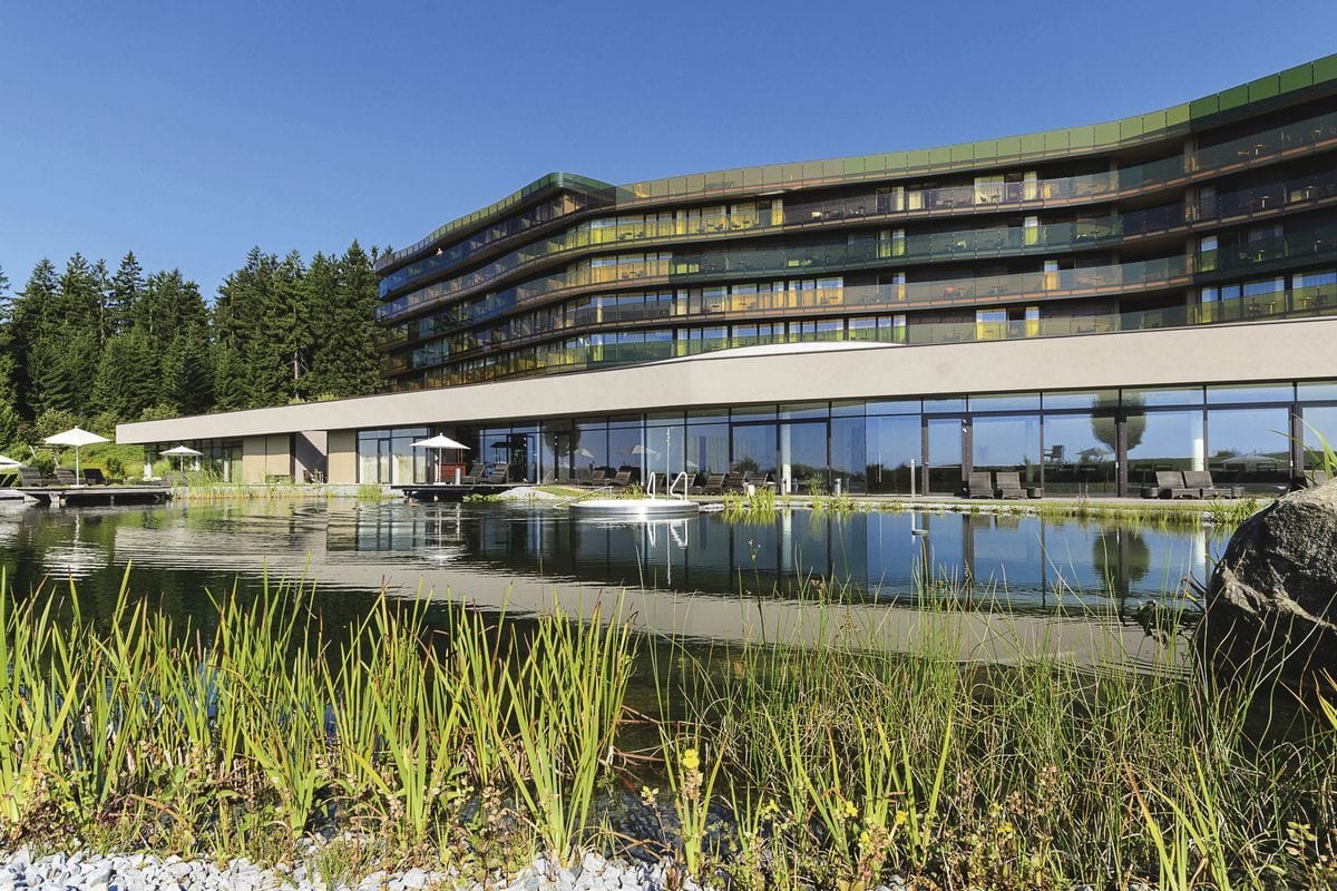 Ein modernes Hotelgebäude mit Glasfassade und Balkonen spiegelt sich in einem naturnahen Schwimmteich mit Schilfbewuchs und Felsen im Vordergrund bei Tag.