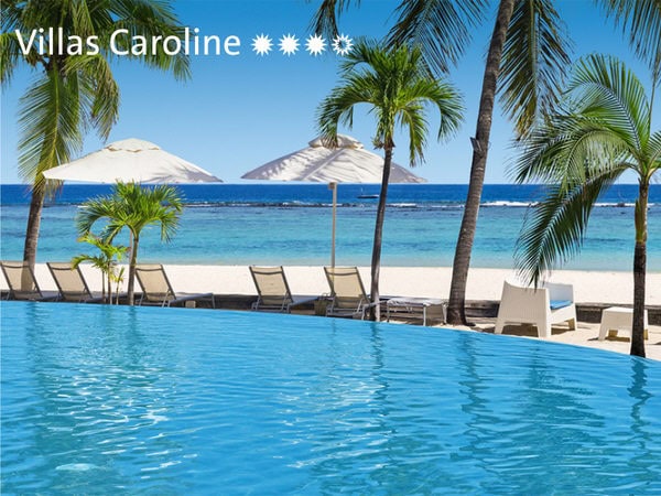 Blick über einen Infinity-Pool auf einen Sandstrand mit Palmen, weißen Sonnenschirmen und Liegestühlen vor dem tiefblauen Meer bei Villas Caroline.