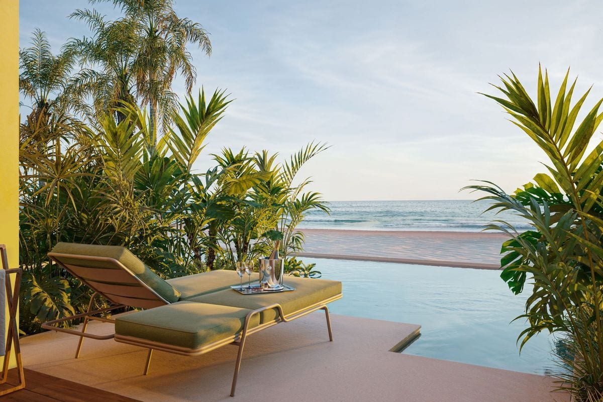 Zwei grüne Sonnenliegen stehen auf einer Terrasse direkt an einem Pool mit Blick auf einen Sandstrand und das Meer, umgeben von üppiger tropischer Vegetation.