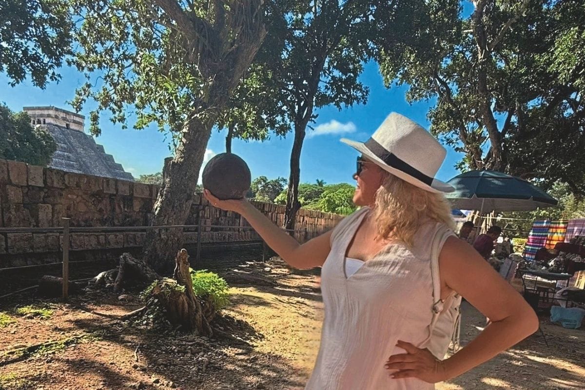 Eine Frau mit Sonnenhut und Sonnenbrille steht vor einer Maya-Pyramide und hält eine dunkle Steinkugel auf ihrer flachen Hand in die Höhe, im Freien.