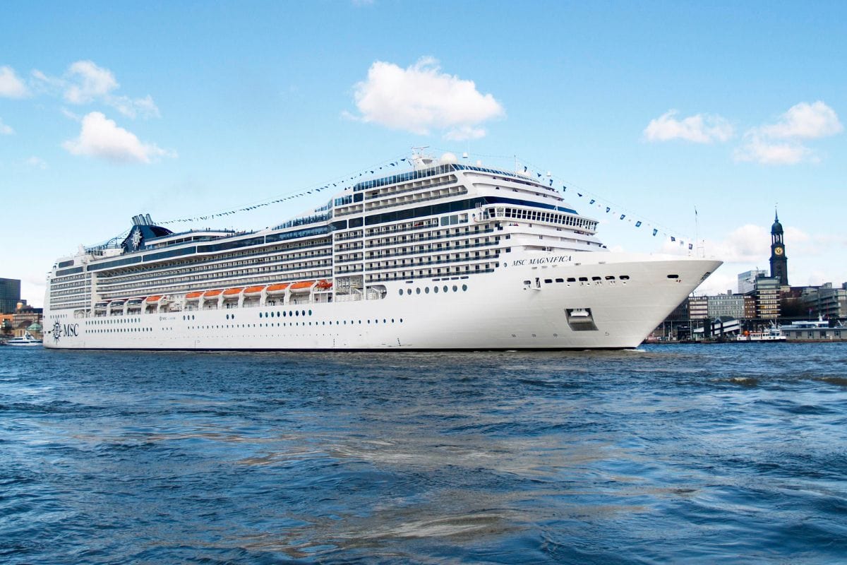Das Kreuzfahrtschiff MSC Magnifica fährt auf dem Wasser vor einer städtischen Kulisse mit einem markanten Kirchturm bei blauem Himmel und Sonnenschein.