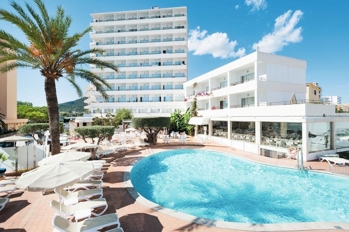 Ein weißes Hotelgebäude mit vielen Balkonen und einem großen Außenpool, umgeben von einer Terrasse mit zahlreichen Sonnenliegen und einer hohen Palme.