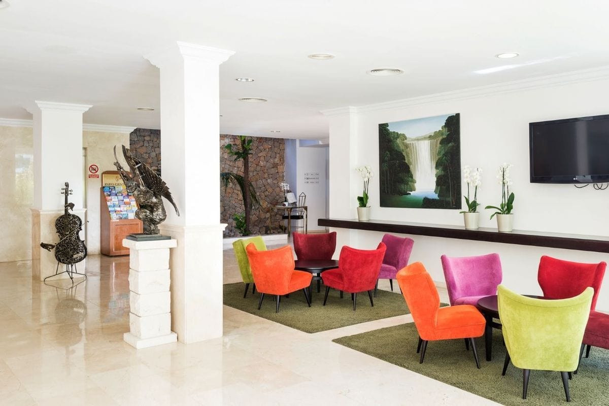 Heller Hotel-Lobbybereich mit hellen Fliesen, weißen Säulen, einer Skulptur und Sitzgruppen aus farbenfrohen Sesseln auf grünen Teppichen vor einer Wand.