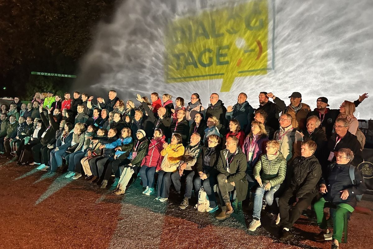 Eine große Gruppe von Menschen sitzt bei Nacht vor einer Wasserfontäne, auf die das gelbe Logo der Dialogtage mit einem Smiley projiziert wird.