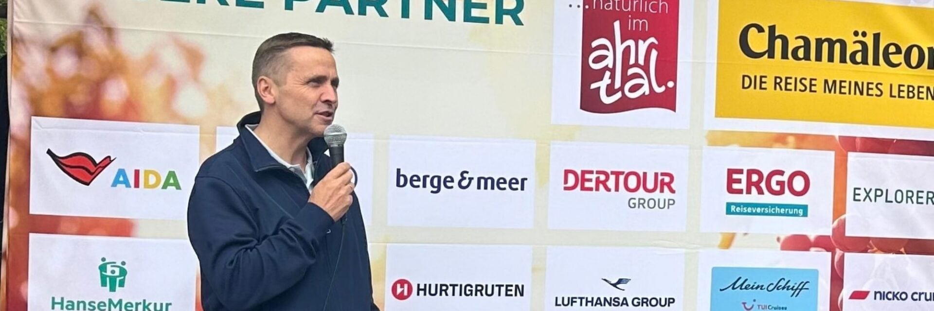 Ein Mann in einer dunkelblauen Jacke spricht vor einer Sponsorenwand mit verschiedenen Firmenlogos wie AIDA, Dertour und HanseMerkur in ein Mikrofon.