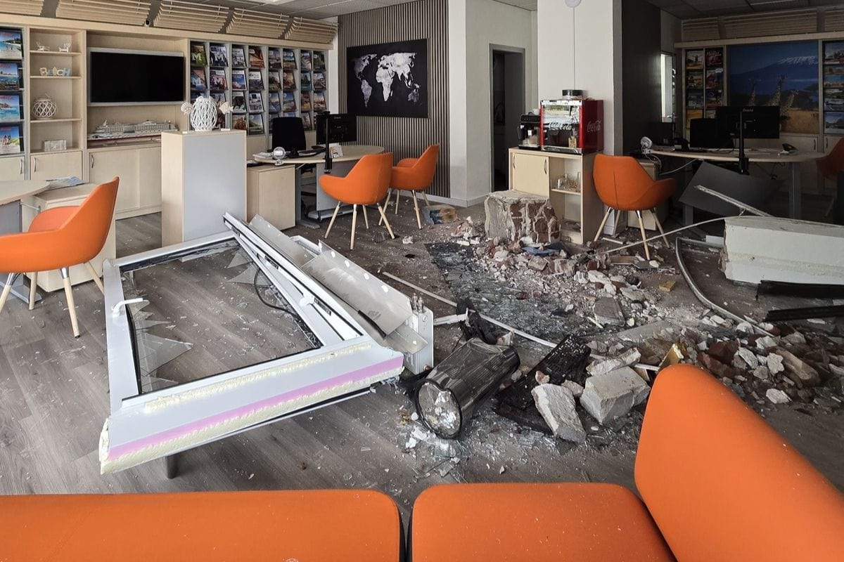 Ein verwüstetes Reisebüro mit einem herausgebrochenen Fensterrahmen, Trümmern, Glassplittern und Ziegelsteinen auf dem Boden zwischen orangefarbenen Stühlen.