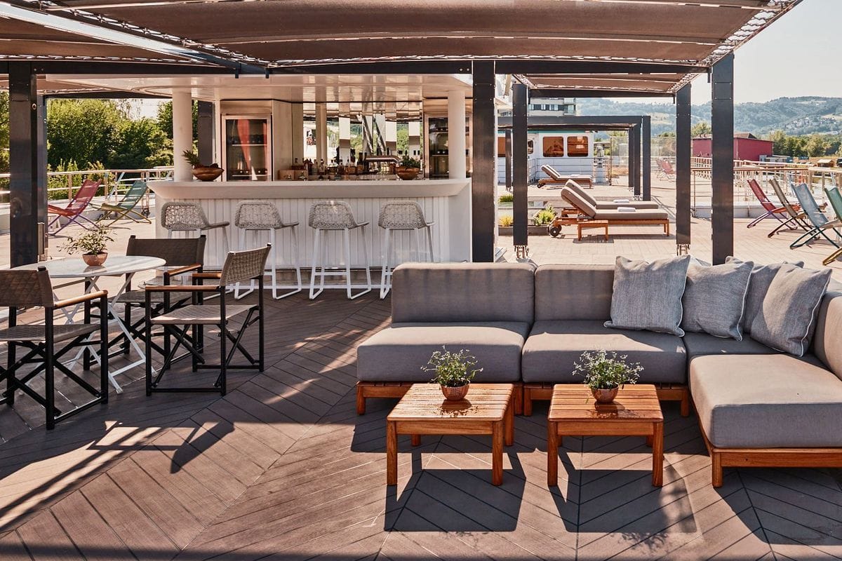 Ein weitläufiges Sonnendeck auf einem Schiff mit einer Bar, Sitzgruppen, Liegestühlen und einem Holzboden unter einer schattenspendenden Überdachung.