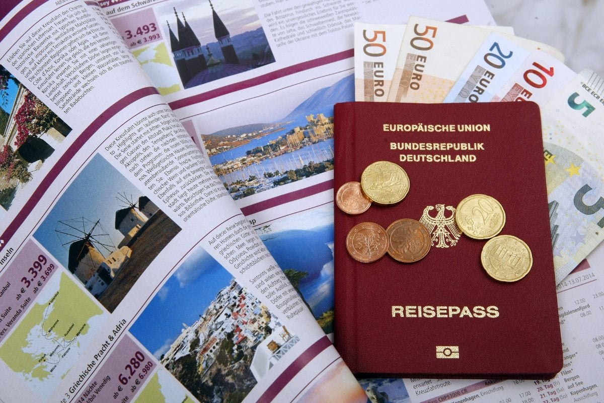 Ein deutscher Reisepass liegt zusammen mit verschiedenen Euro-Banknoten und Münzen auf einer aufgeschlagenen Reisebroschüre mit Bildern von Urlaubsregionen.