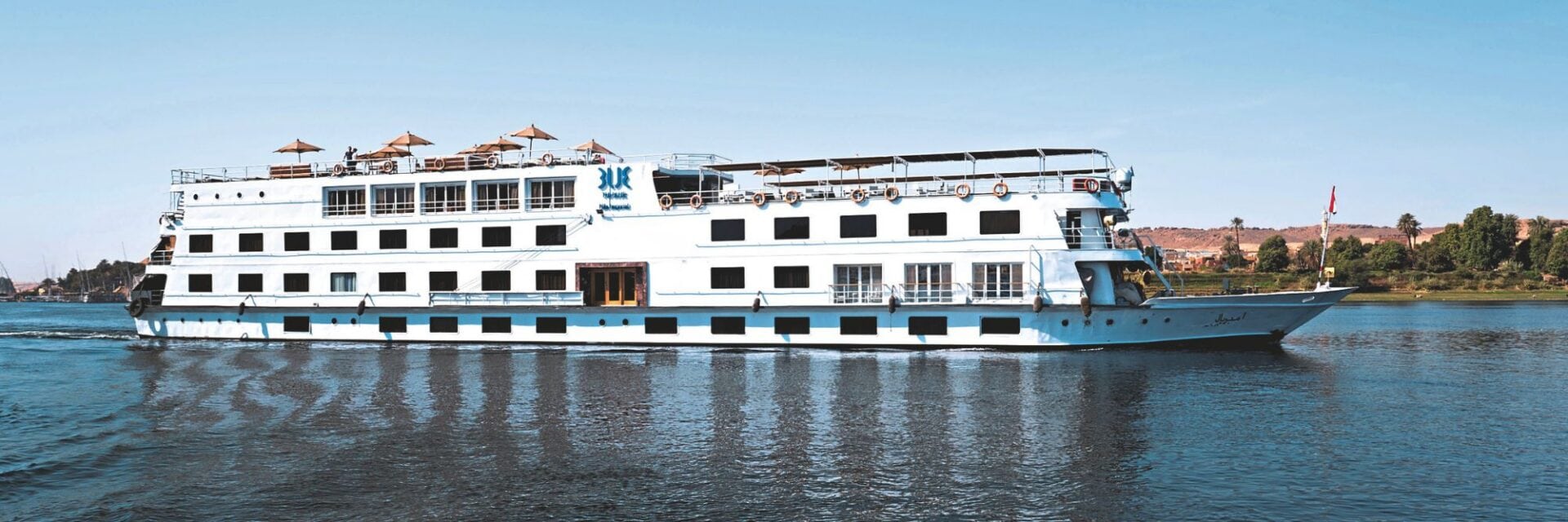 tui-nil-kreuzfahrt Ein weißes Nilkreuzfahrtschiff fährt auf dem ruhigen Wasser des Nils unter einem klaren blauen Himmel, mit Sonnenschirmen auf dem obersten Deck zu sehen.