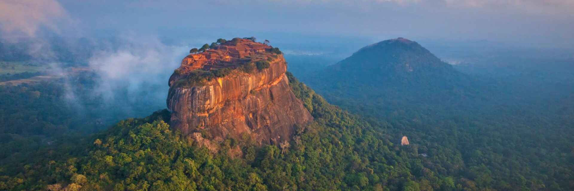 Website Header - 2026-10-14T112027.715 Luftaufnahme des Sigiriya-Felsens in Sri Lanka, der aus einem dichten, nebligen Dschungel emporragt, mit einem weiteren bewaldeten Hügel im Hintergrund.