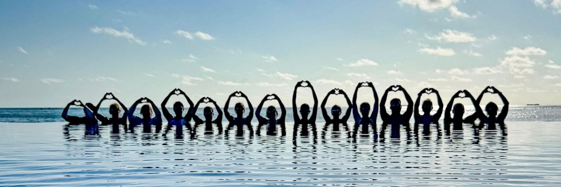 Eine Reihe von Personen steht bis zur Brust im Wasser eines Infinity-Pools vor dem Meer und formt mit den Händen Herzsymbole gegen den hellen Himmel.