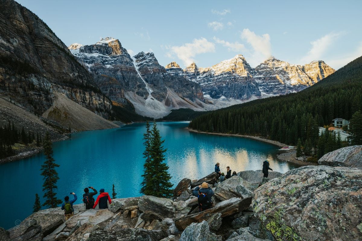 Eine Gruppe von Menschen steht auf felsigem Untergrund und blickt auf den türkisfarbenen Moraine Lake, umrahmt von schneebedeckten Bergen und Nadelwäldern.