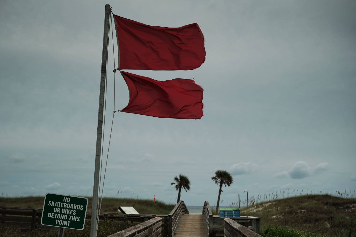 Zwei rote Flaggen wehen an einem Mast vor einem bewölkten Himmel über einem Holzsteg, der zu einem Strand mit Dünen und zwei Palmen im Hintergrund führt.