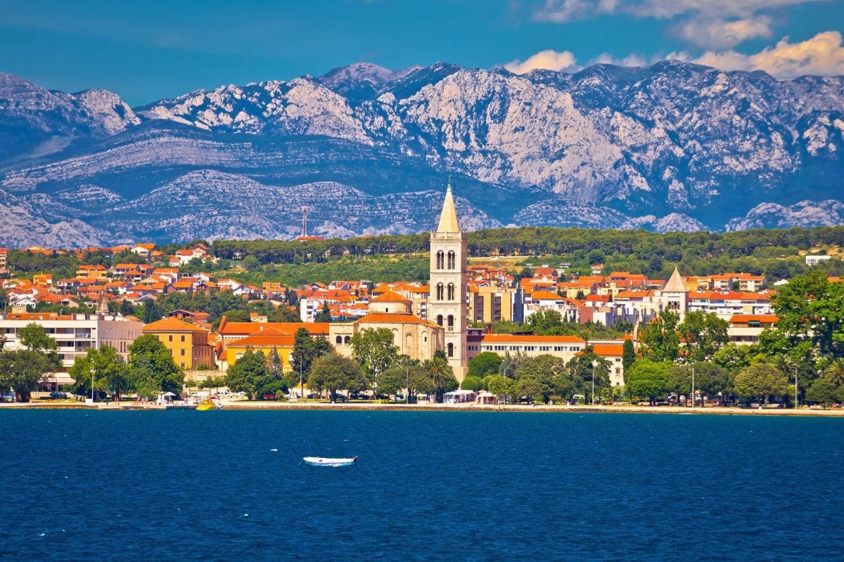 Blick auf eine kroatische Küstenstadt mit Kirchturm und roten Dächern vor einem massiven, schroffen Gebirgsmassiv unter einem klaren, blauen Sommerhimmel.
