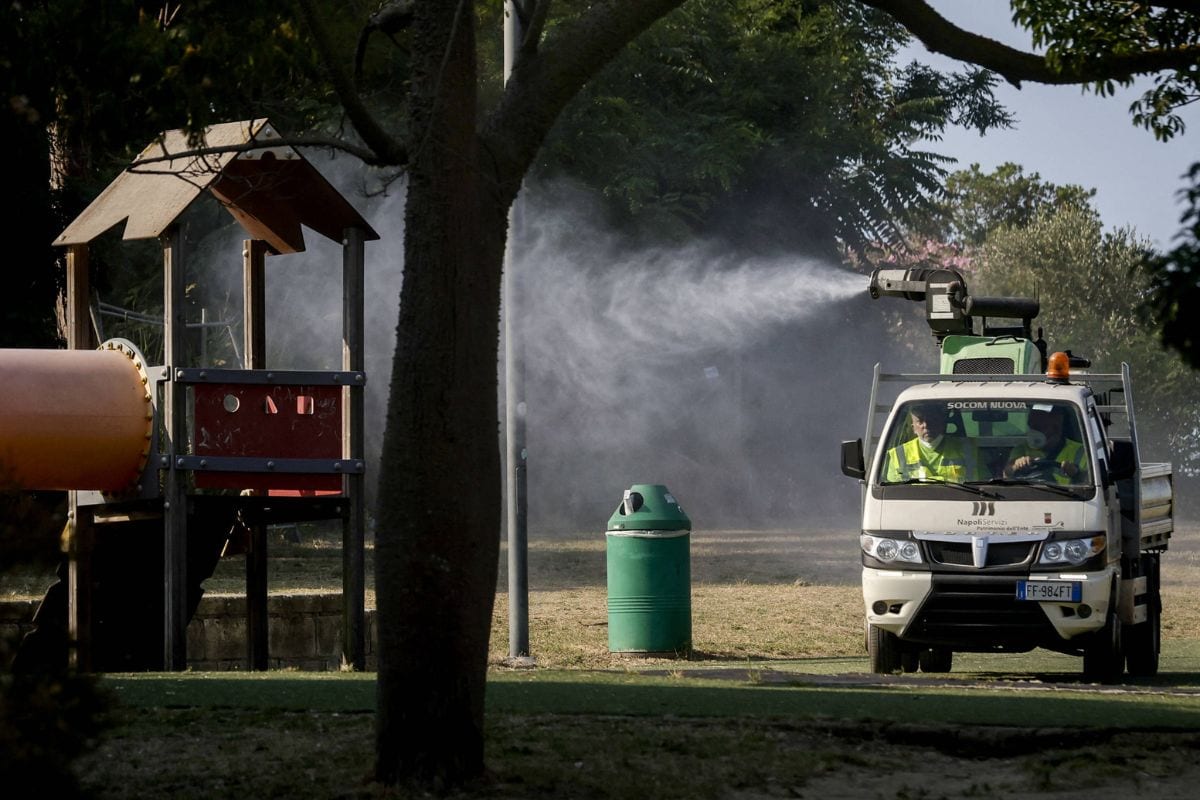 Ein weißer LKW mit Sprühvorrichtung fährt an einem Spielplatz vorbei und verteilt einen feinen Nebel in der Luft, während ein Mülleimer im Vordergrund steht.
