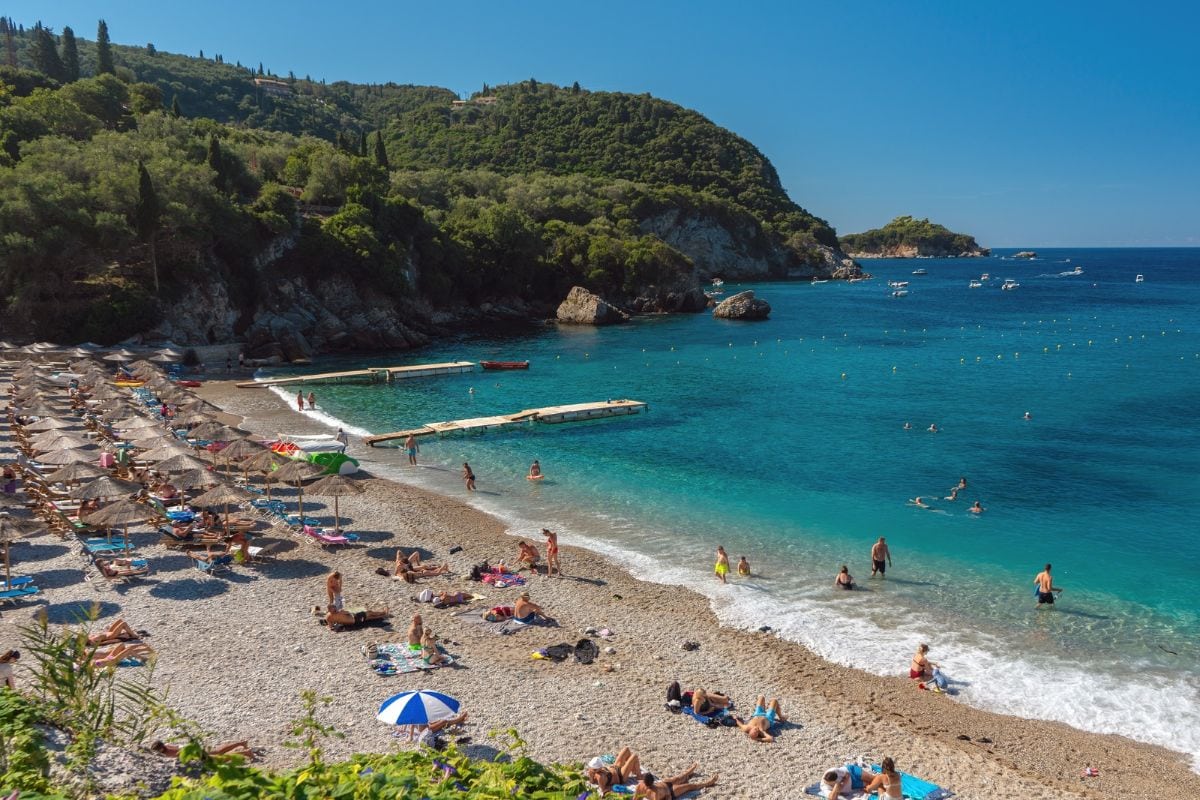 Blick auf einen Kieselstrand mit zahlreichen Sonnenschirmen, badenden Menschen im türkisfarbenen Wasser und bewaldeten Hügeln im Hintergrund bei Sonnenschein.