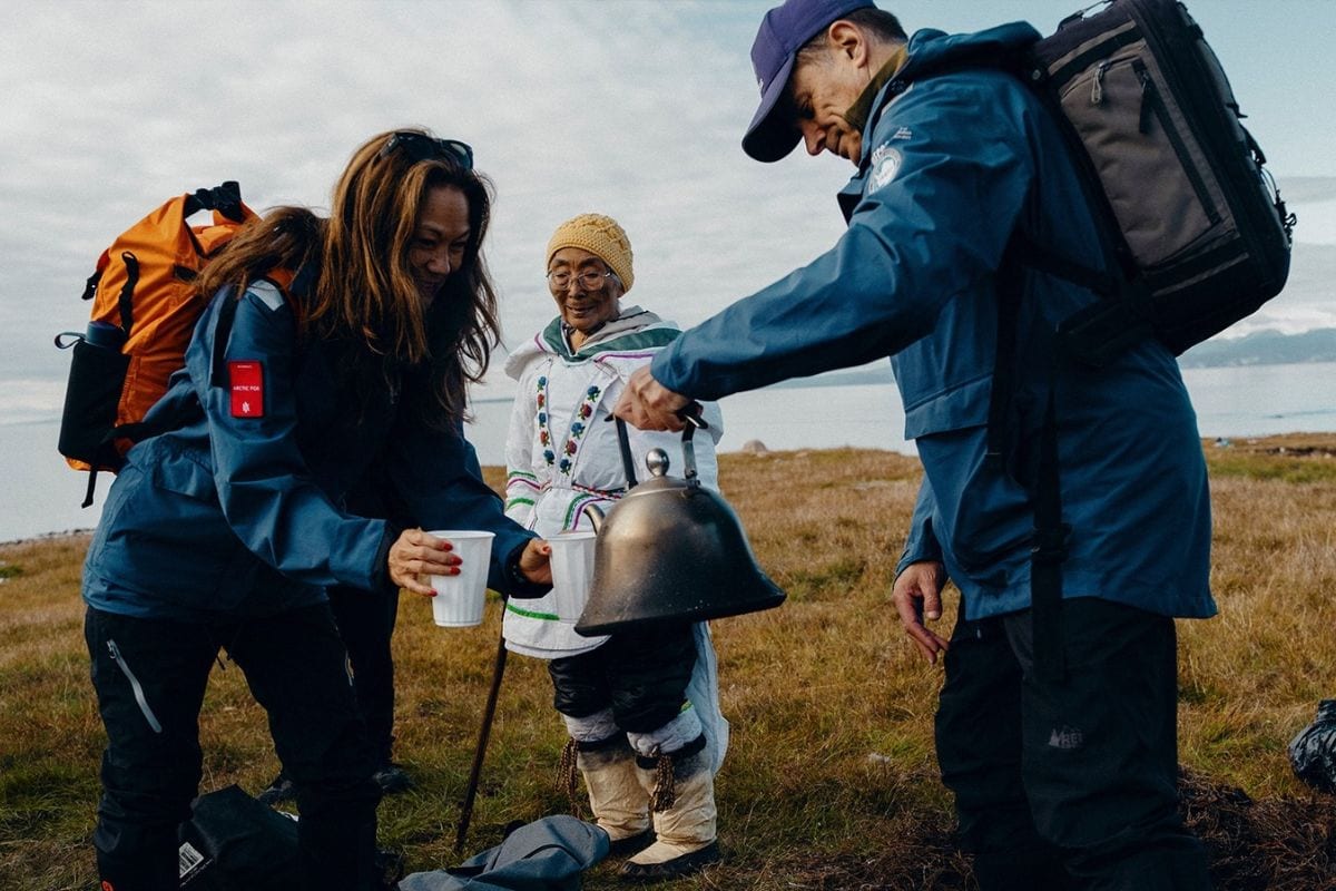 Zwei Personen in Outdoor-Kleidung schenken einer älteren Frau in traditioneller Kleidung mit einer Metallkanne ein heißes Getränk in Becher ein.