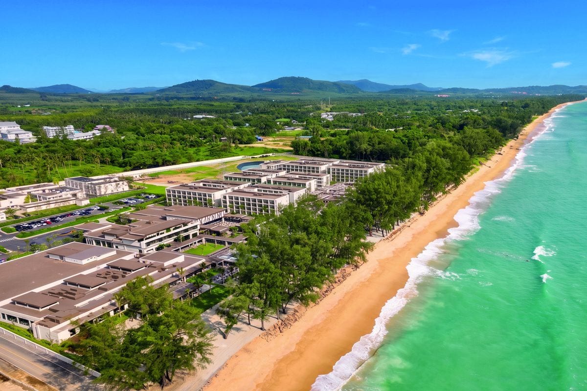 Luftaufnahme eines großen Hotelkomplexes direkt an einem langen Sandstrand mit türkisfarbenem Meer, umgeben von grünen Bäumen und einer hügeligen Landschaft.