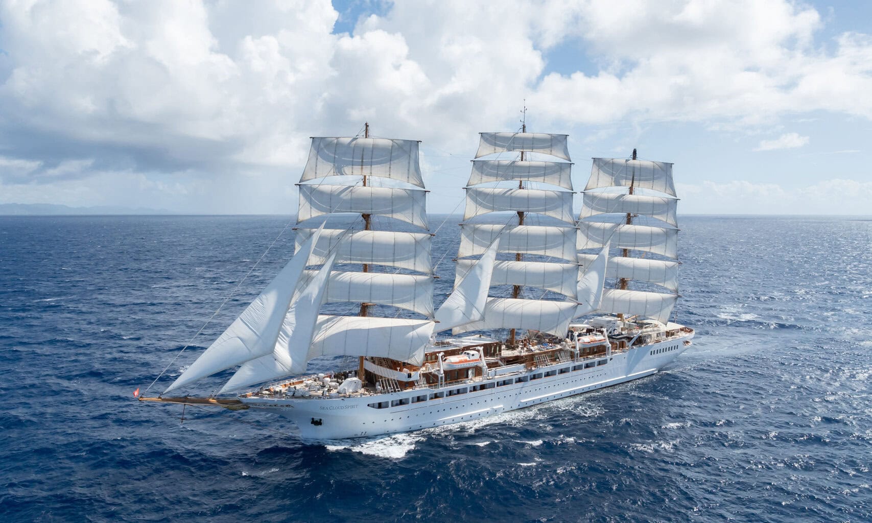 Die Sea Cloud Spirit, ein weißer Dreimast-Segler mit gesetzten Segeln, fährt bei sonnigem Wetter und leicht bewölktem Himmel über das tiefblaue offene Meer.