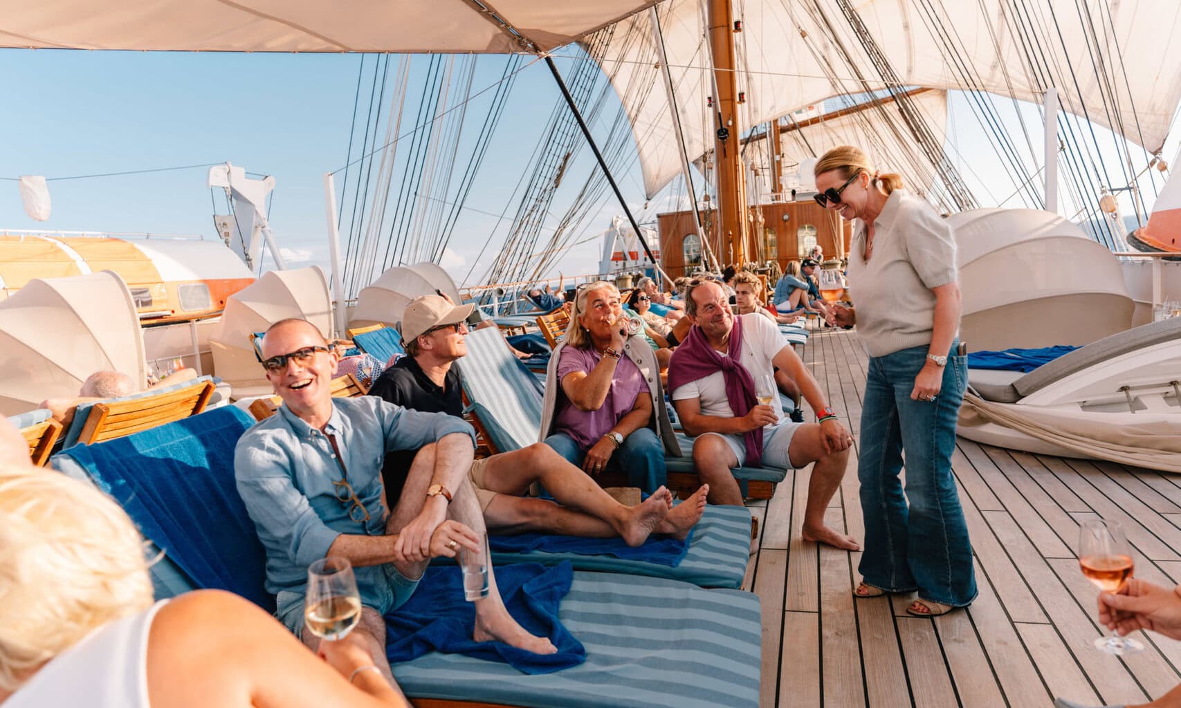 Eine Gruppe von Menschen entspannt sich auf dem Holzdeck eines Segelschiffs in Liegestühlen, während sie Getränke hält und sich untereinander unterhält.