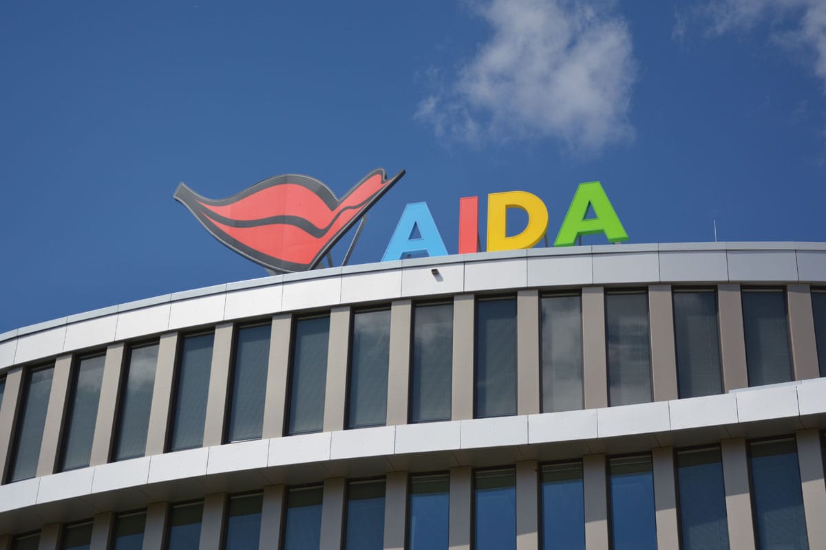Das bunte AIDA-Logo mit dem charakteristischen Kussmund auf dem Dach eines modernen Bürogebäudes mit Glasfassade vor einem klaren blauen Himmel mit Wolken.