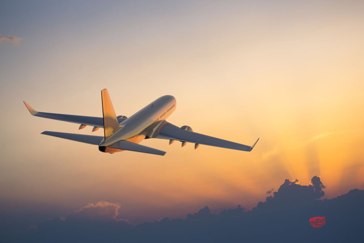 Ein Passagierflugzeug fliegt schräg von hinten gesehen in den goldenen Abendhimmel über einer Wolkendecke, während die Sonne am Horizont tief steht.