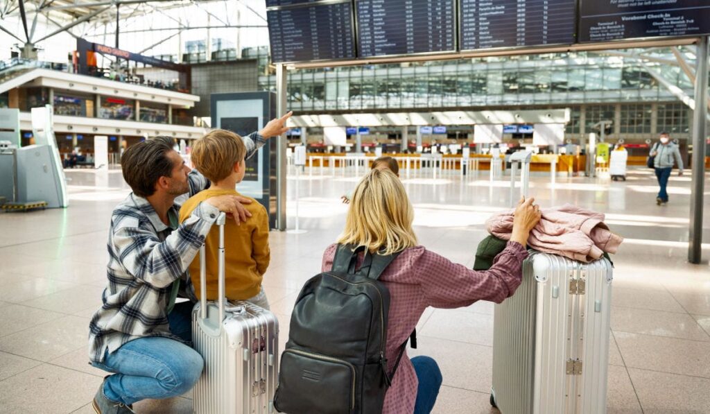 Eine vierköpfige Familie kniet in einer hellen Flughafenhalle vor einer großen Anzeigetafel für Abflüge, während der Vater auf einen Flugbildschirm deutet.