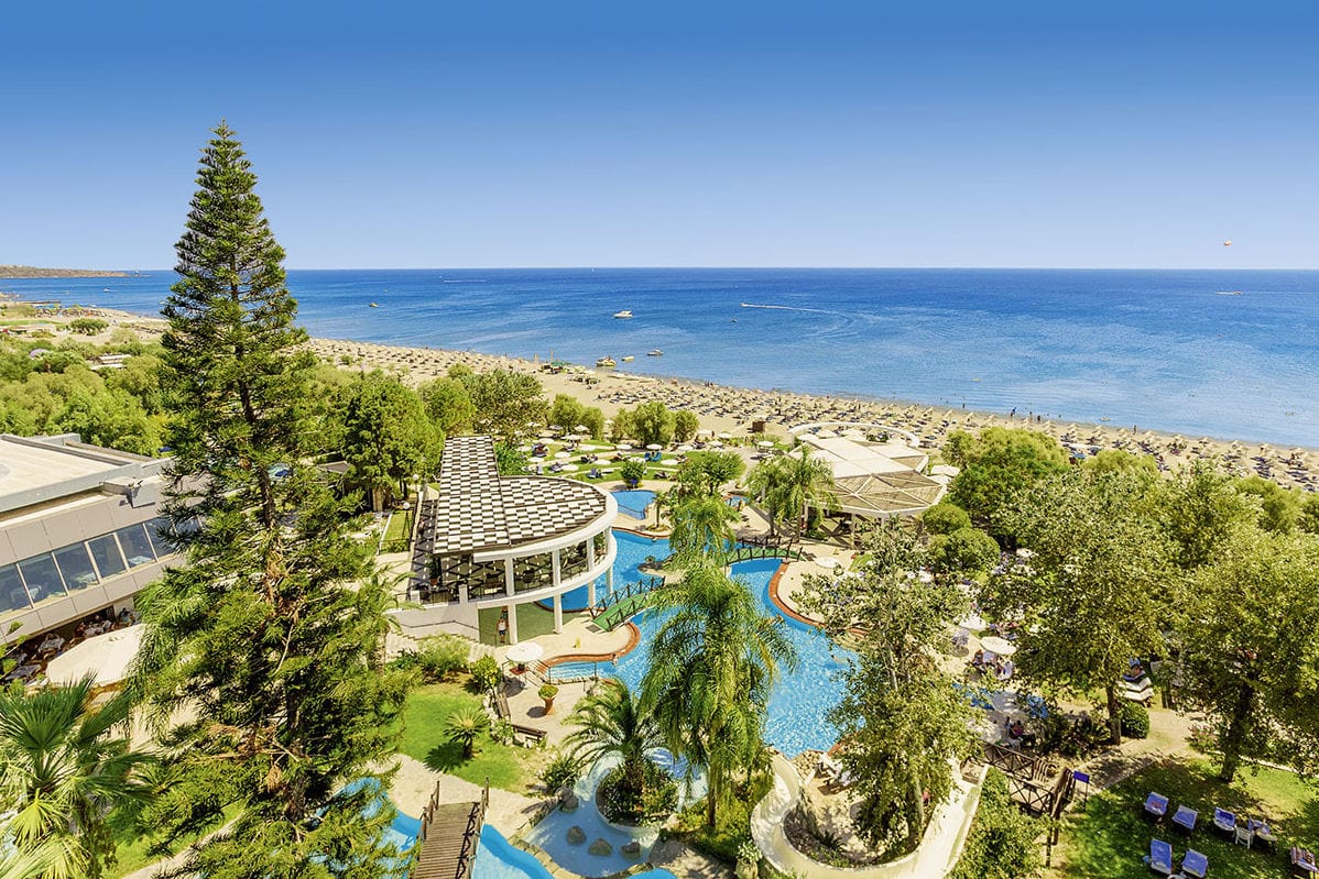 Blick von oben auf eine Hotelanlage mit Poollandschaft, Palmen und Grünflächen, die direkt an einen breiten Sandstrand und das offene Meer angrenzt.