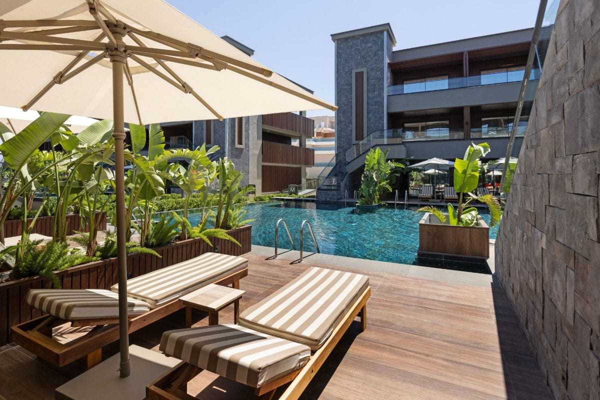 Zwei gestreifte Sonnenliegen auf einer Holzterrasse vor einem Hotelpool mit Palmen und einem großen Sonnenschirm bei strahlendem Sonnenschein im Außenbereich.