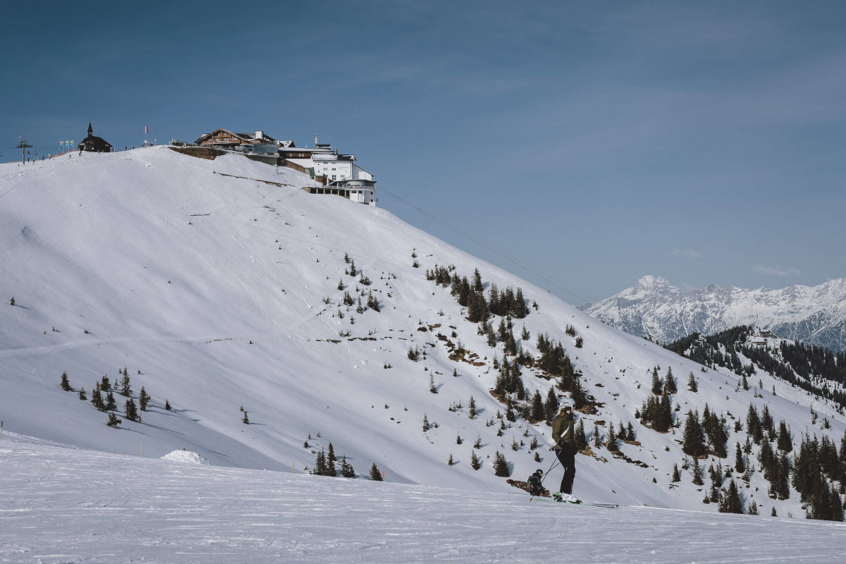 Eine Skifahrerin steht auf einer verschneiten Piste vor einem Berghang, auf dessen Gipfel sich ein großes Hotelgebäude unter blauem Himmel befindet.