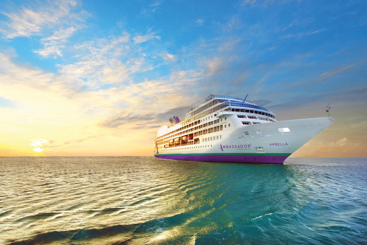 Das Kreuzfahrtschiff Ambella der Reederei Ambassador fährt bei Sonnenuntergang auf dem offenen Meer unter einem wolkenverhangenen, hellen Abendhimmel.