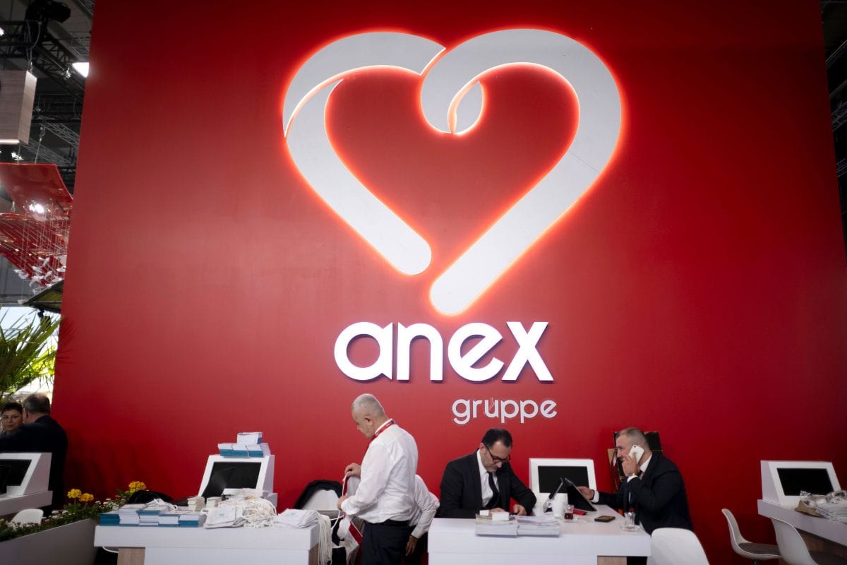 Ein roter Messestand mit dem beleuchteten Logo der anex gruppe, an dem drei Männer an weißen Schreibtischen arbeiten und einer von ihnen telefoniert.