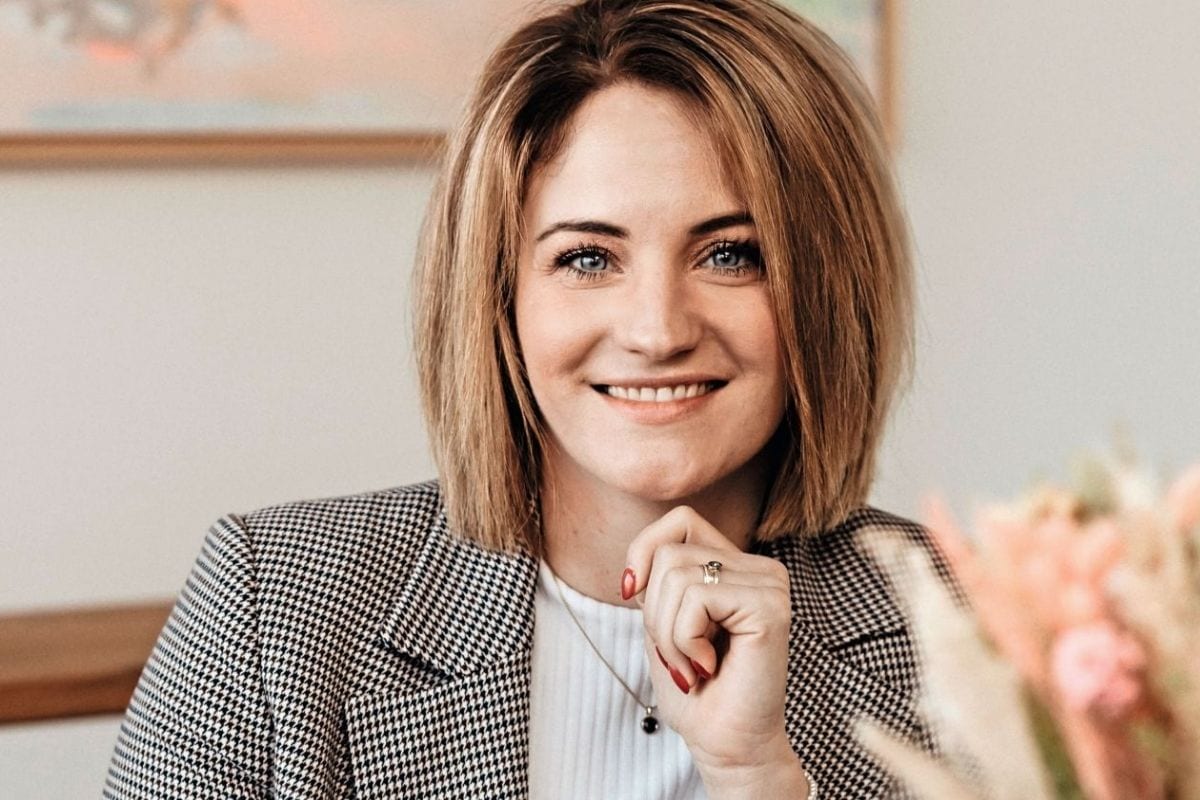 Eine Frau mit schulterlangem, blondem Haar und einem karierten Blazer lächelt freundlich in die Kamera, während sie ihre Hand unter das Kinn hält.