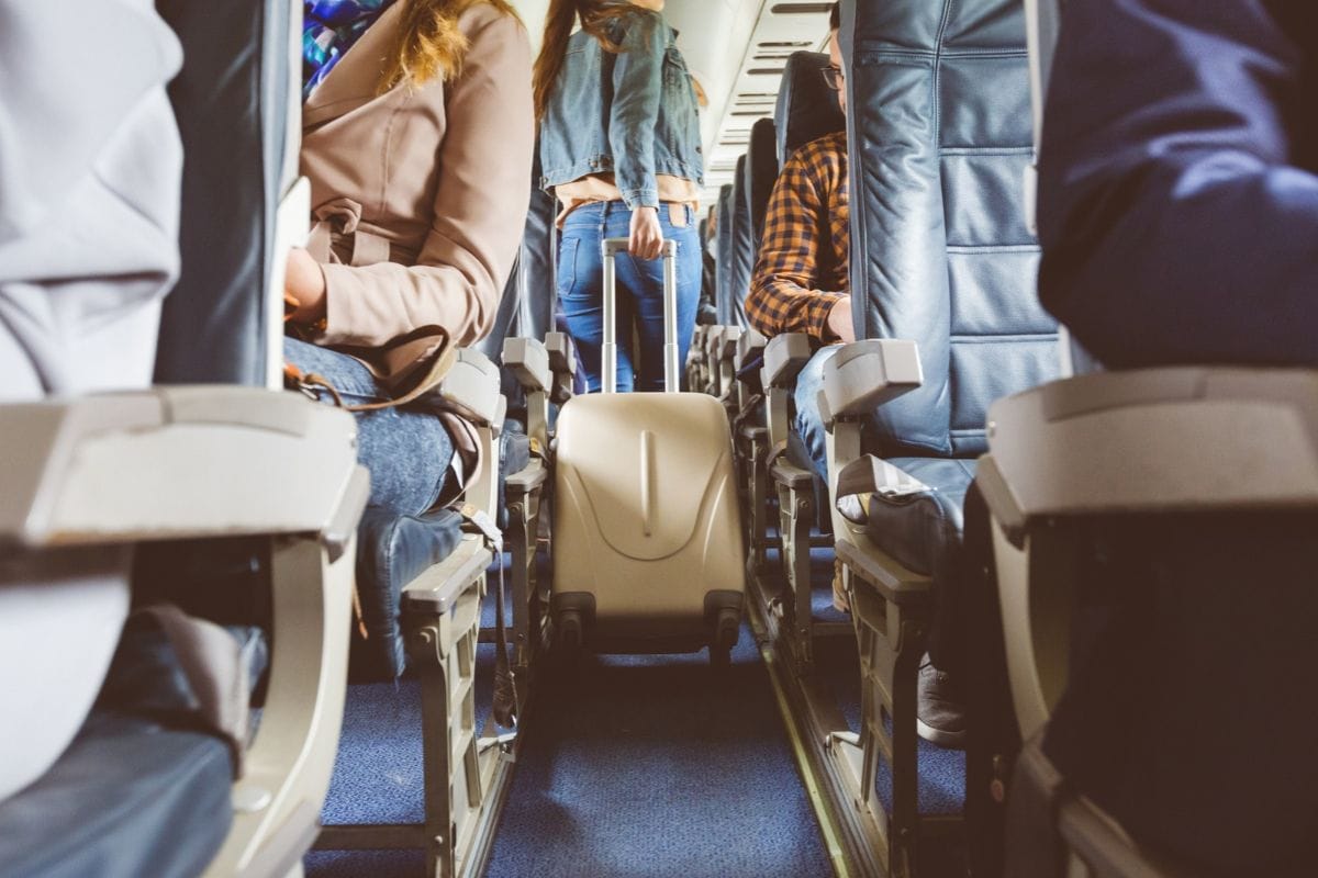 Blick durch den Mittelgang eines Flugzeugs mit Passagieren auf den Sitzen und einer Person, die einen hellen Rollkoffer in den Gang schiebt, von hinten.