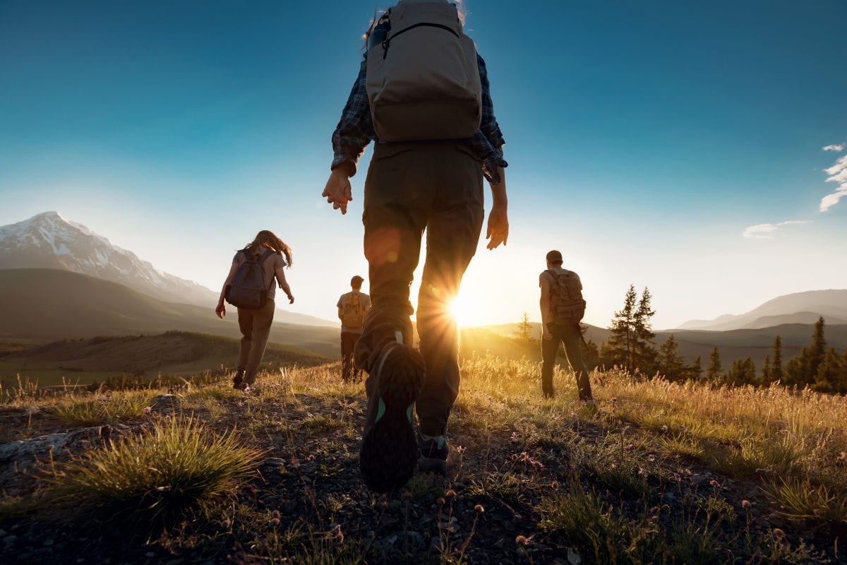 Vier Personen mit Rucksäcken wandern in einer bergigen Landschaft bei Sonnenuntergang auf einem Pfad durch hohes Gras in Richtung der tiefstehenden Sonne.