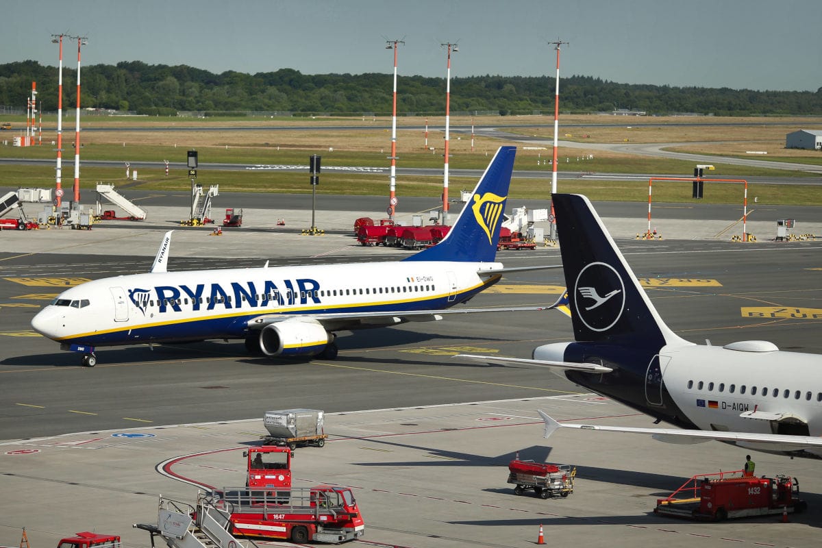 Ein Ryanair-Flugzeug rollt auf dem Rollfeld an einer parkenden Lufthansa-Maschine vorbei, umgeben von Bodenabfertigungsfahrzeugen auf einem Flughafen.