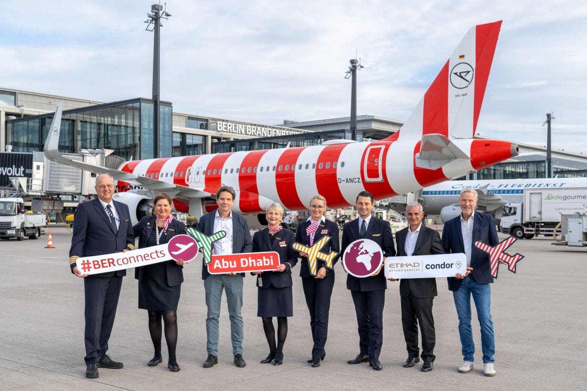 Eine Gruppe von acht Personen steht vor einem rot-weiß gestreiften Flugzeug am Flughafen Berlin Brandenburg und hält verschiedene Schilder in die Kamera.