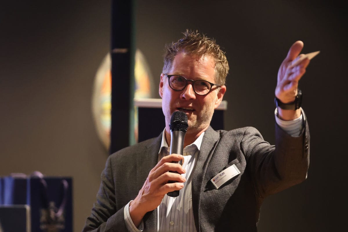 Ein Mann mit Brille und grauem Sakko spricht in ein Mikrofon und hebt dabei gestikulierend seine linke Hand vor einem dunklen Hintergrund im Innenraum.