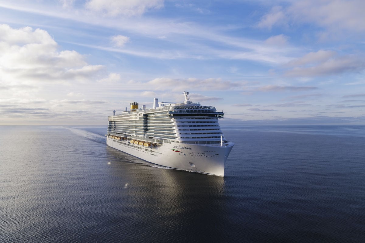 Das Kreuzfahrtschiff Costa Toscana fährt auf dem offenen Meer unter einem blauen Himmel mit leichten Wolkenformationen bei ruhigem Wellengang in der Sonne.
