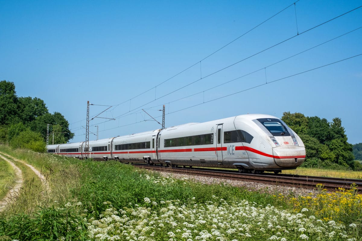 Ein weißer ICE der Deutschen Bahn fährt auf einer zweigleisigen Strecke durch eine grüne Landschaft mit Wiesen und Bäumen unter einem klaren blauen Himmel.