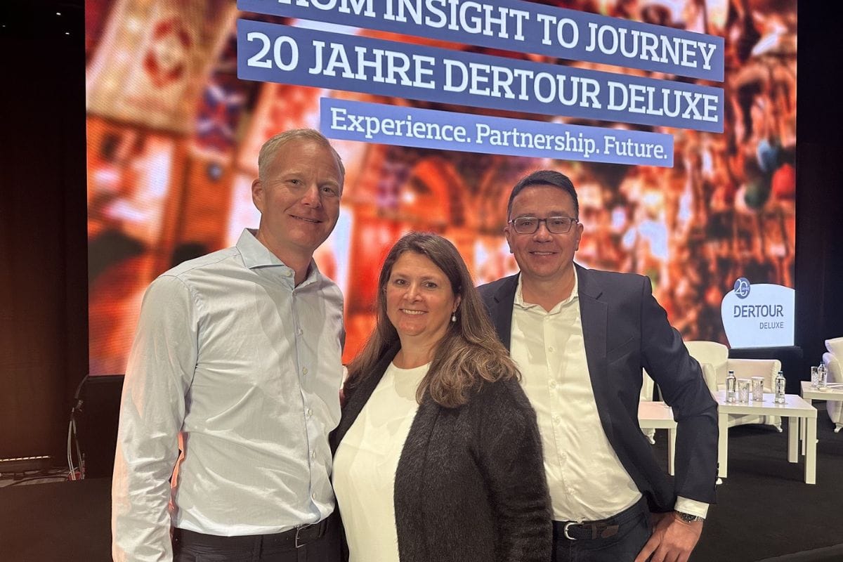 Drei Personen stehen nebeneinander vor einer großen Leinwand mit der Aufschrift 20 Jahre DERTOUR DELUXE und lächeln in die Kamera bei einer Veranstaltung.