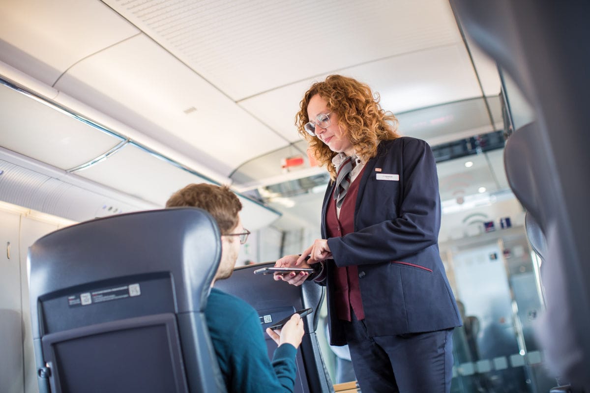 Eine Frau mit rotem Haar und Brille in Uniform steht in einem Zugabteil und zeigt auf ein Smartphone, das ein sitzender Mann in den Händen hält.