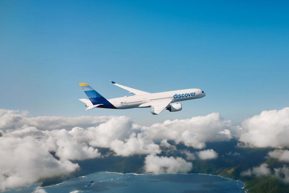 Ein weißes Passagierflugzeug der Fluggesellschaft discover fliegt in Reiseflughöhe über einer Küstenlandschaft mit Wolkenfeldern bei klarem, blauem Himmel.